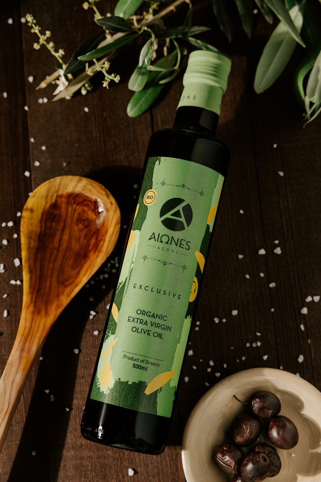 Bottle of AEONS Organic Extra Virgin Olive Oil on a wooden surface with olives and a wooden spoon.